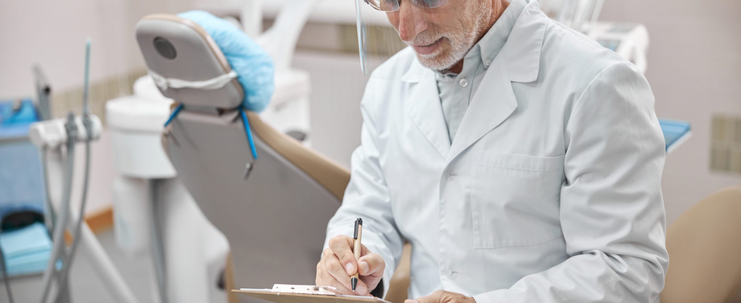 Elderly dental specialist wearing a full-face shield looking down on his notes while sitting in his clinic